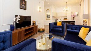 A modern living room with a large flat-screen TV, blue sofas with yellow cushions, a wooden entertainment unit, and a chandelier.