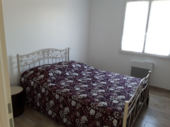A single bed with a floral bedspread in a room with a window, a radiator, and a small round table. - Fully equipped villa with swimming pool (Limoux)