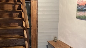 Una escalera de madera que conduce a una puerta, un banco y un tapete estampado en una habitación con suelo de baldosas y un cuadro colgado en la pared.