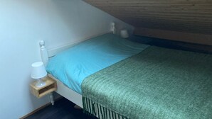 Una habitación pequeña con cama individual, techo inclinado, tragaluz, mesita de noche con lámpara y una colcha verde.