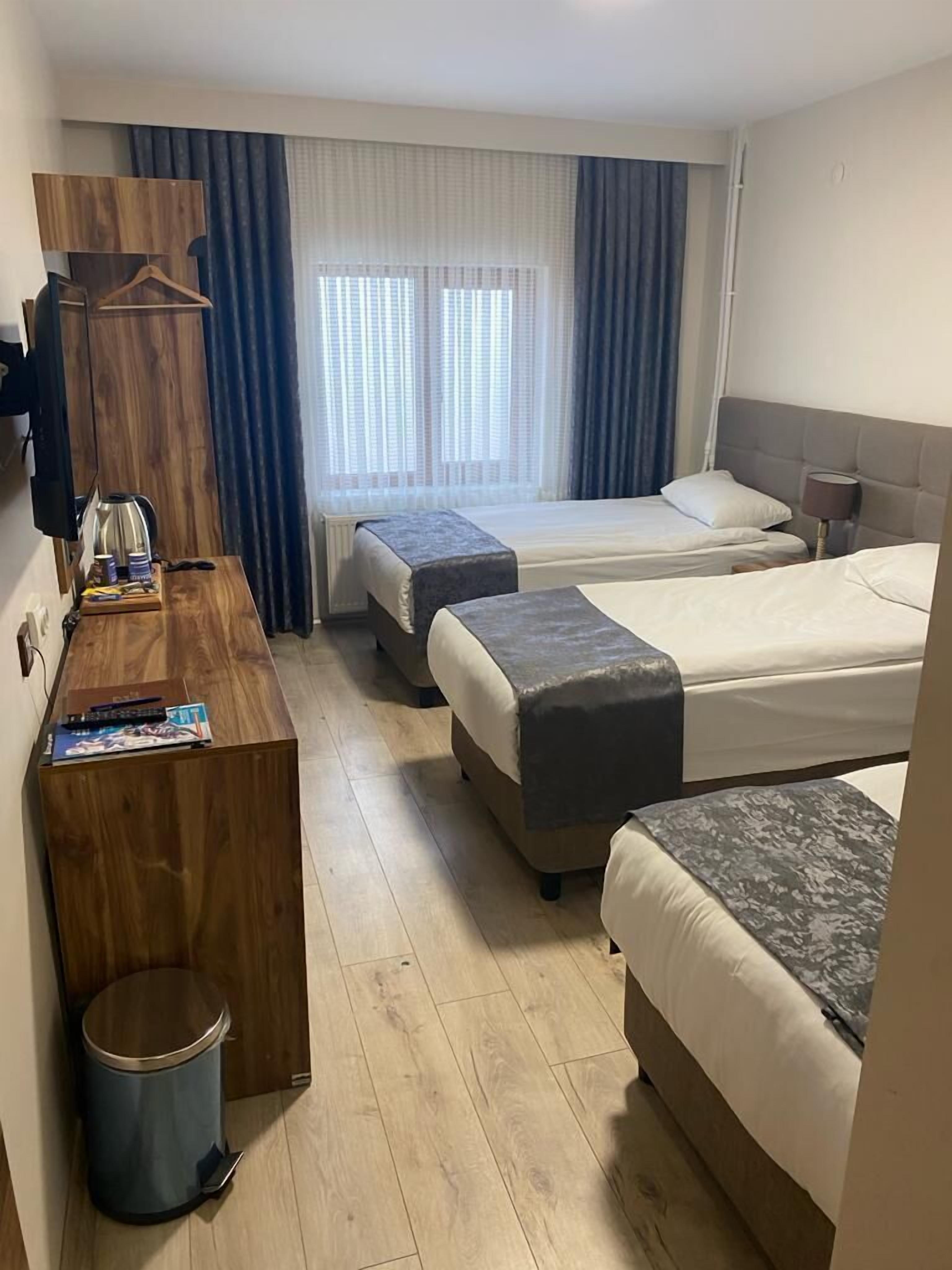 Une chambre d’hôtel avec deux lits simples, un bureau en bois, une poubelle et une fenêtre avec des rideaux.