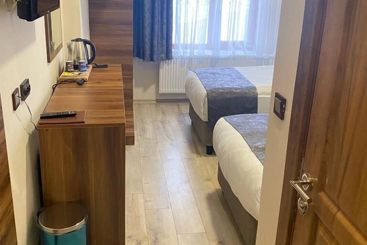 A compact hotel room with a wooden desk, a small trash bin, a bed, and a window with curtains.