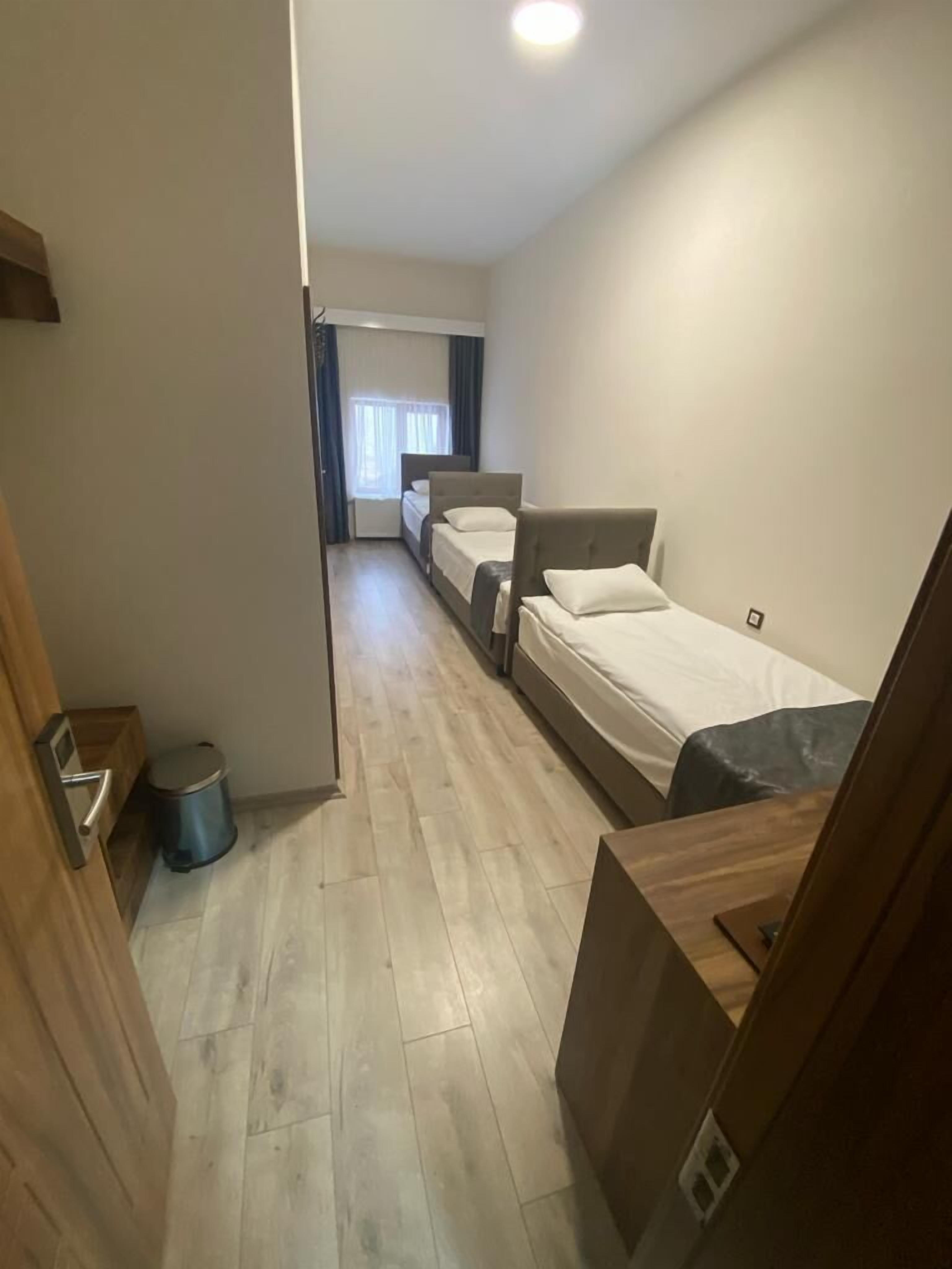 A narrow hallway leading to a room with two beds, a trash bin, and a wooden door.