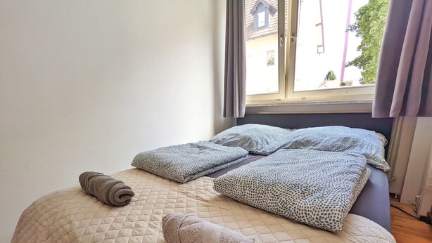 A neatly made bed with a quilted headboard and footboard, a window with curtains, and a view of a building outside. - Vacation Apartment Near The Black Forest (Villingen-Schwenningen)