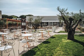 An outdoor seating area with white tables and chairs, a pergola, and a large tree.