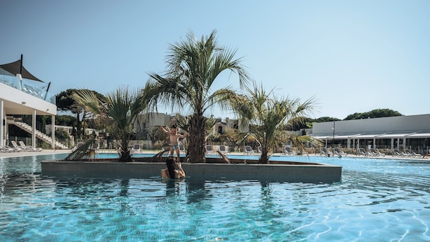 A swimming pool with palm trees, lounge chairs, and a modern building in the background. - Belambra Clubs Port Camargue - Les Salins (Le Grau-Du-Roi)