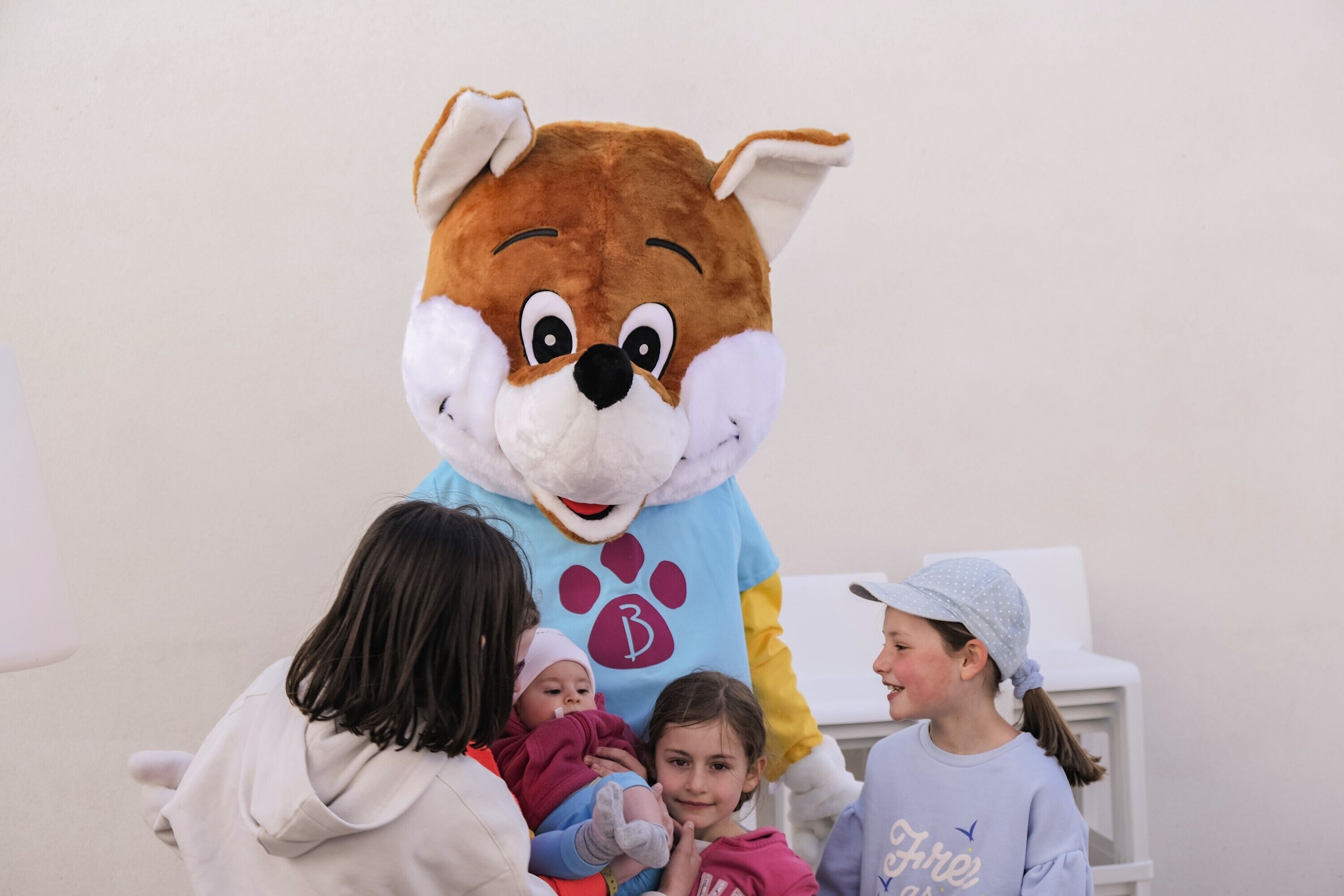 Une mascotte interagissant avec des enfants et des adultes dans une pièce dont le mur et la chaise sont blancs.