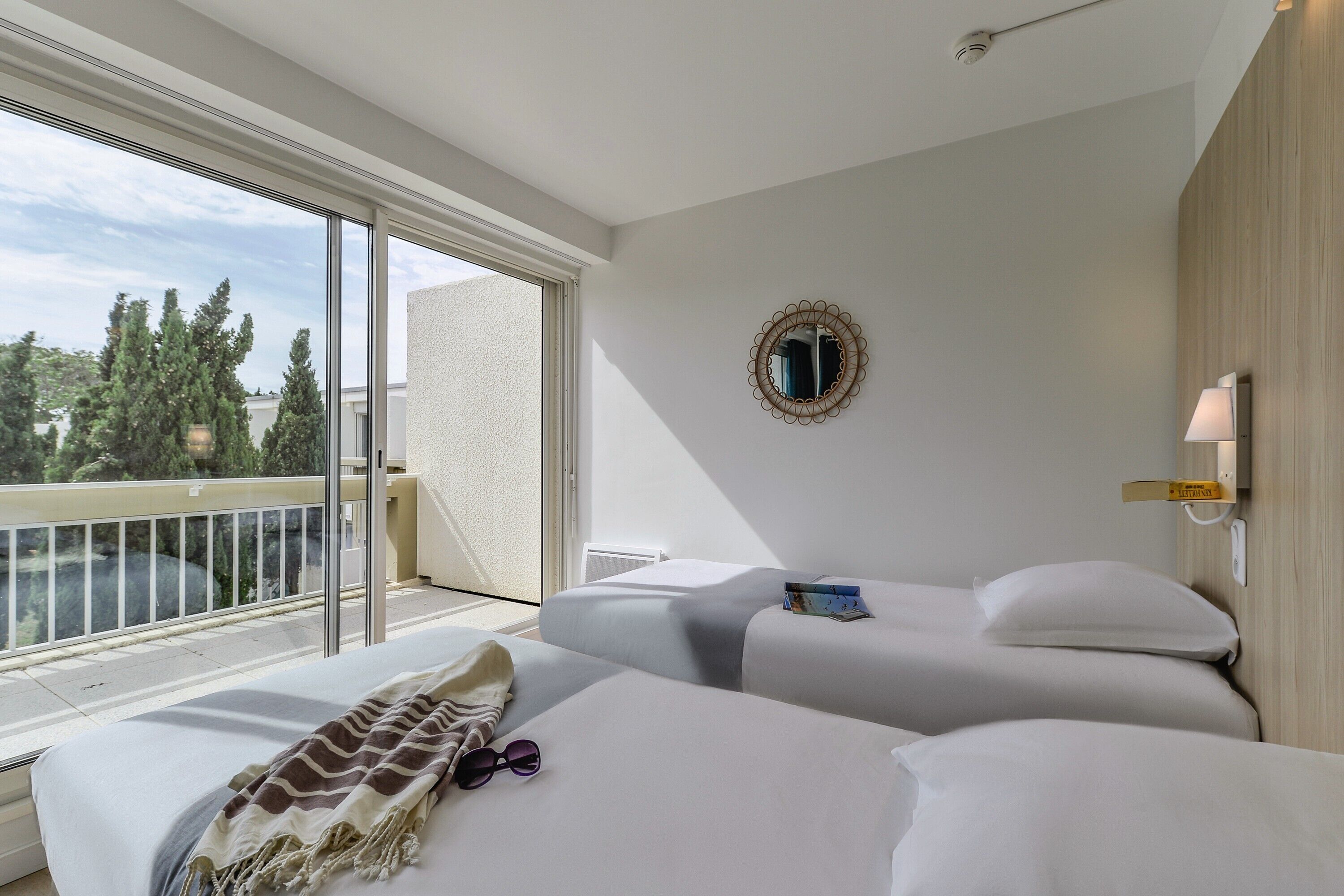 Habitación de hotel con dos camas, un ventanal con vistas a árboles, un espejo redondo en la pared y una lámpara de noche.