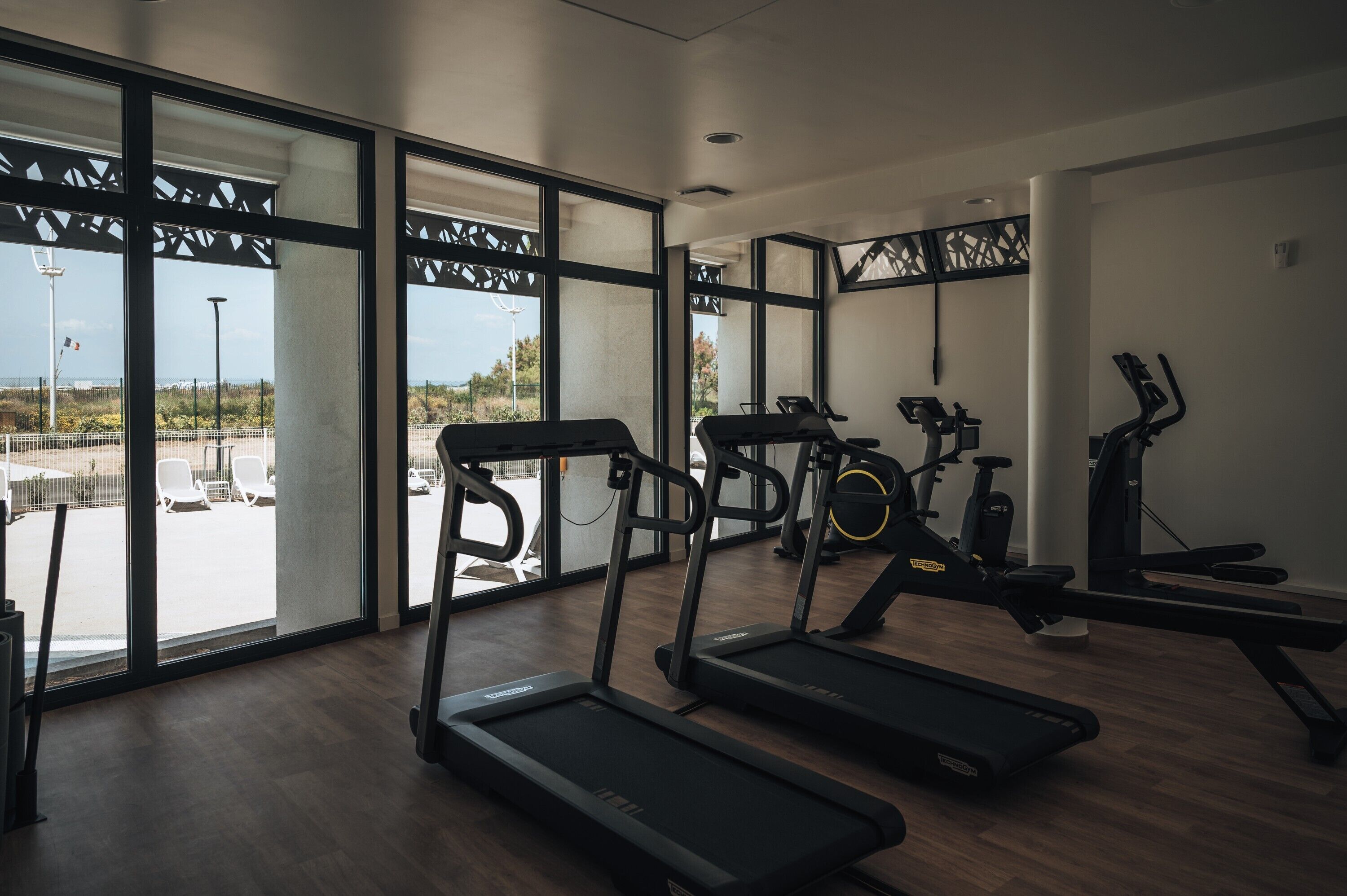 Un gimnasio con cintas de correr, bicicletas estáticas y máquinas de remo. Amplias ventanas ofrecen vistas al exterior.