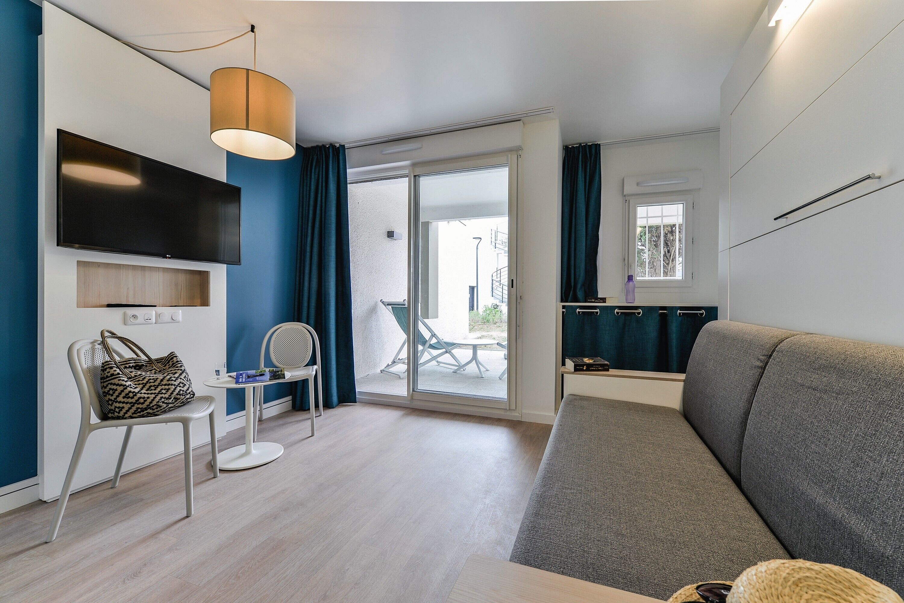 Un salon moderne avec un canapé gris, un téléviseur à écran plat fixé au mur, une petite table ronde avec une chaise et une porte-fenêtre coulissante donnant sur un espace extérieur.