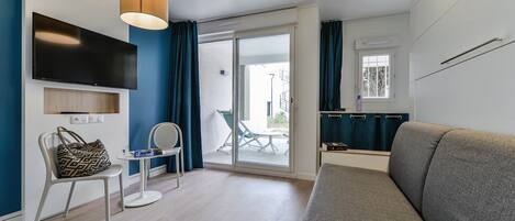 Un salon moderne avec un canapé gris, un téléviseur à écran plat fixé au mur, une petite table ronde avec une chaise et une porte-fenêtre coulissante donnant sur un espace extérieur.