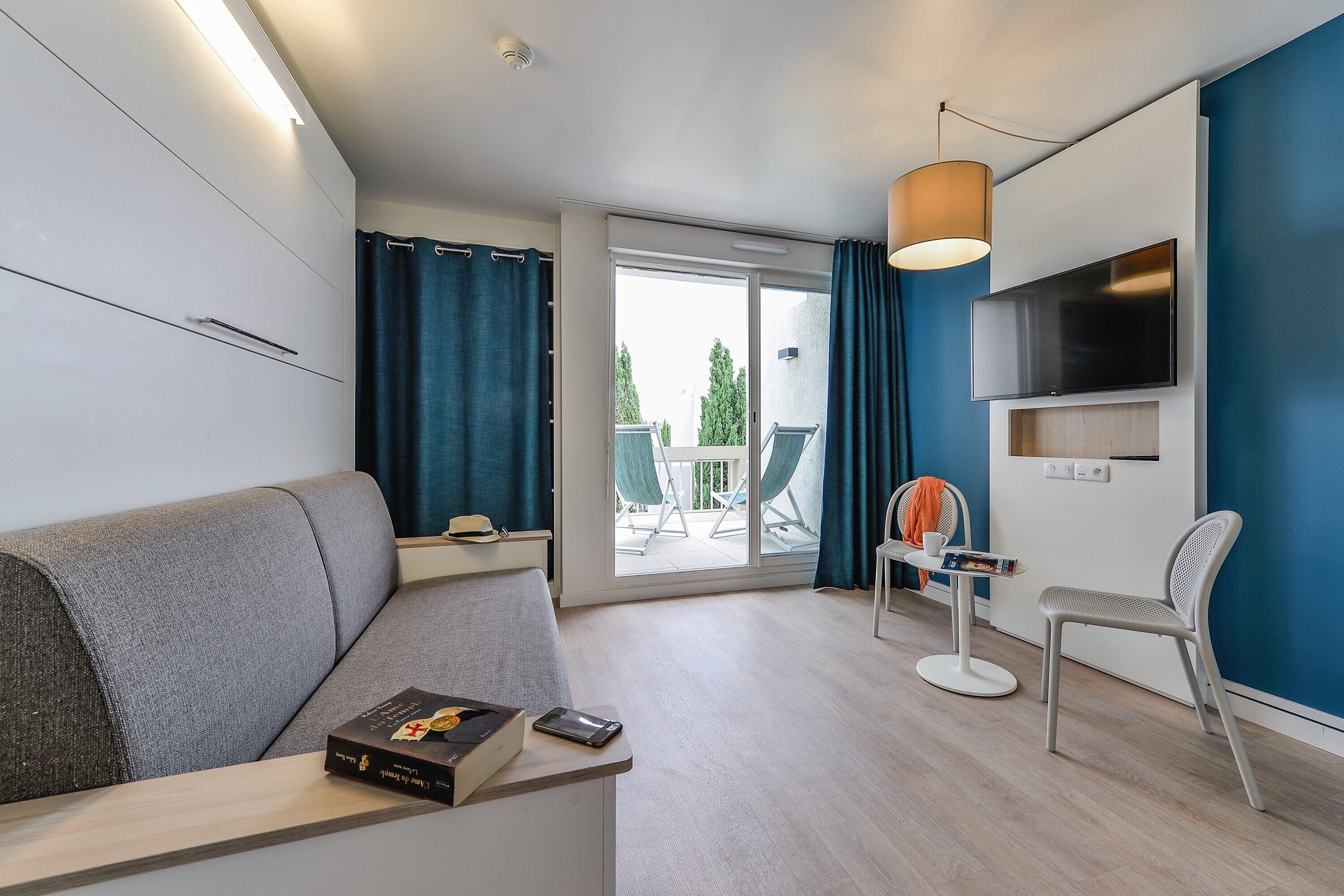 Een moderne hotelkamer met een bank, een klein tafeltje met een boek en een smartphone, een aan de muur bevestigde televisie, en een balkon met een tafel en stoelen.