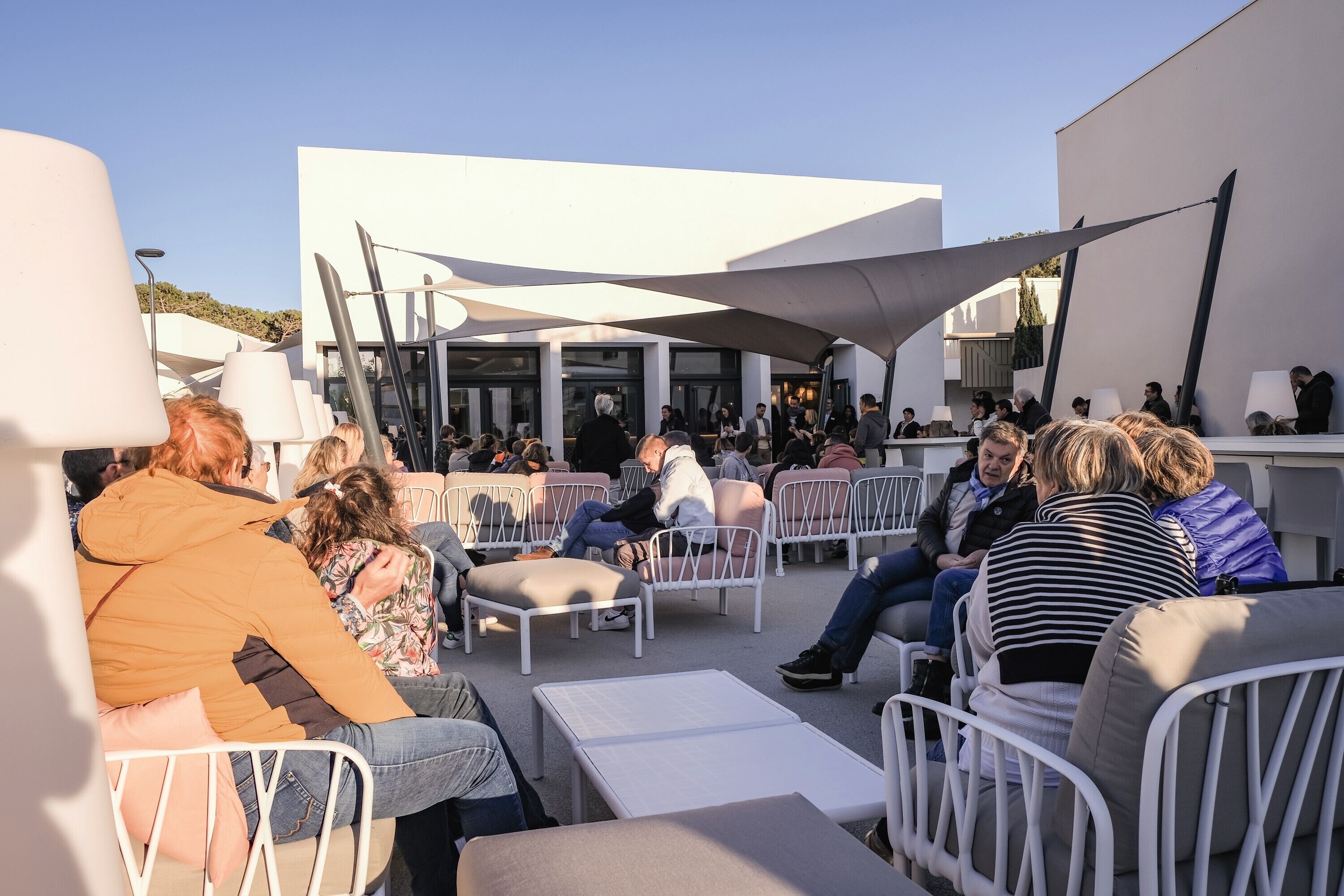 An outdoor seating area with white metal chairs and tables, surrounded by white walls and large umbrellas providing shade.