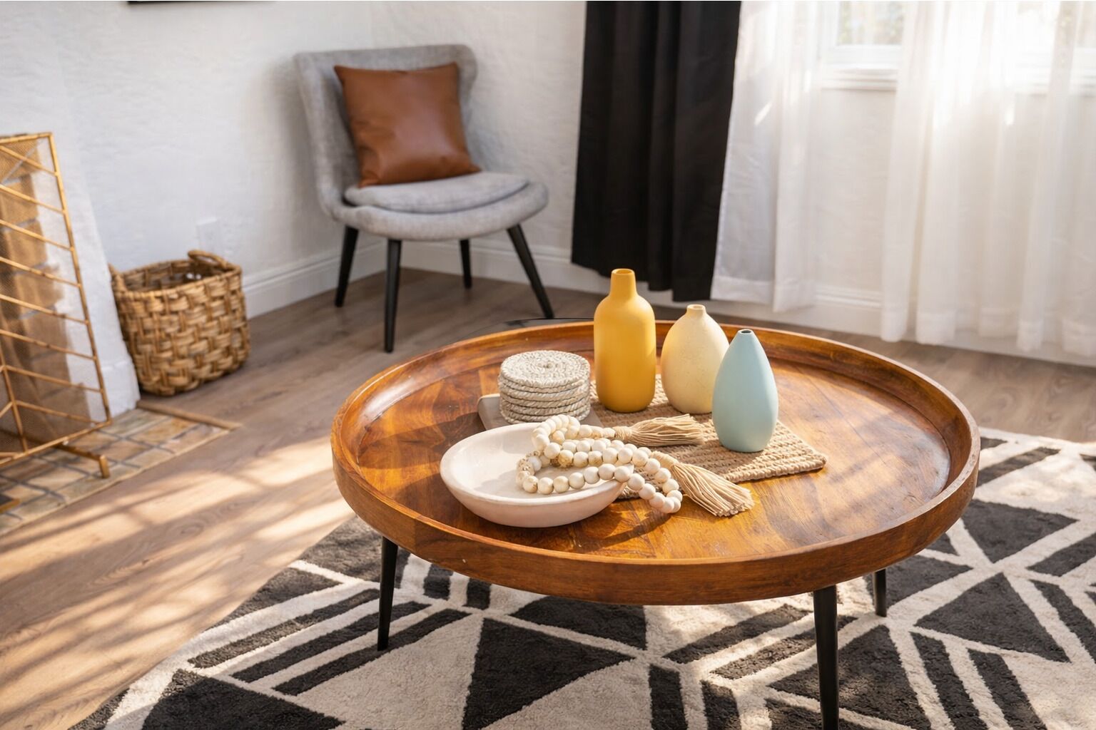 Ein runder Couchtisch aus Holz mit einer gewebten Unterlage, einem Teller mit weißen Perlen und drei Vasen in Gelb, Weiß und Blau.