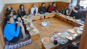 Un groupe de personnes est assis autour d’une table en bois, dans une pièce aux murs et aux bancs également en bois.