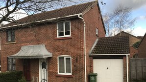 A two-story brick house with a white garage door, a small porch with a metal roof, and a green waste bin labeled "Garden Waste Collection."