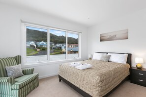 Ein Schlafzimmer mit großem Fenster, einem Bett mit gemustertem Überwurf, einem Nachttisch mit Lampe und einem Sessel mit Streifenmuster auf den Kissen.