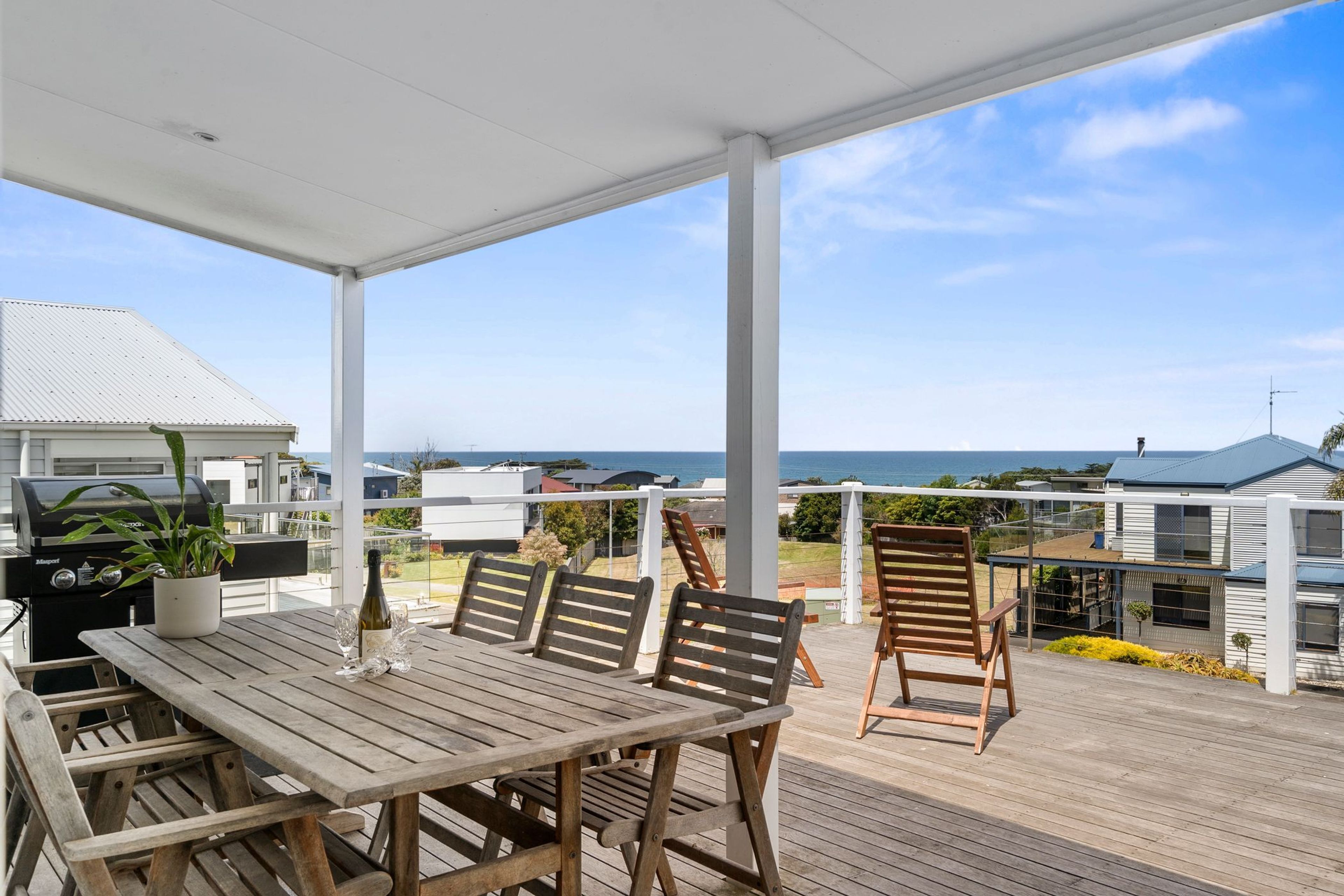 A covered outdoor patio with a wooden table and chairs, overlooking a residential area and the sea.