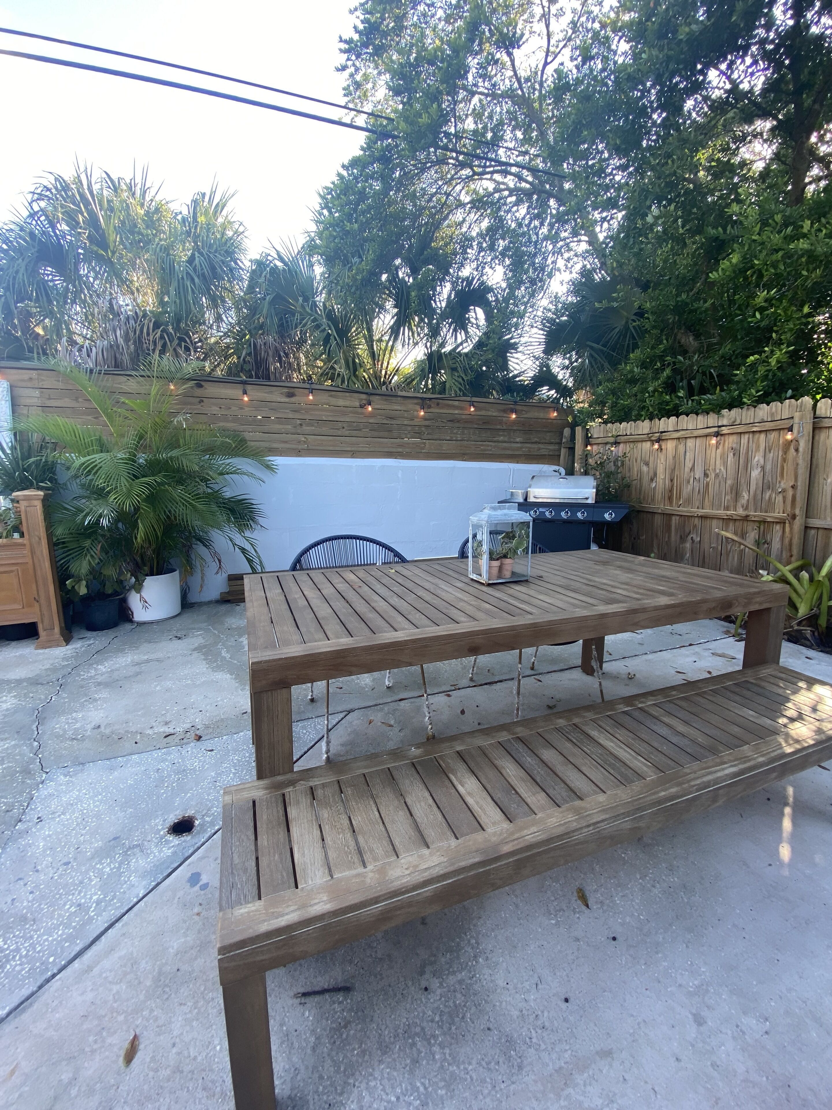 Una mesa de patio de madera con un banco, rodeada de plantas en macetas y una valla de madera.