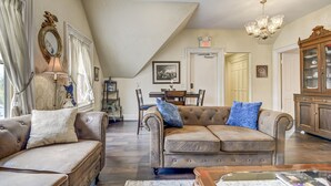 A spacious living room with a high ceiling, a chandelier, a brown leather sofa with blue cushions, a wooden coffee table, and a dining area in the background.