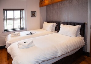 Une chambre d’hôtel avec deux lits, une fenêtre avec des stores, un tableau encadré au mur et une poutre en bois au plafond.