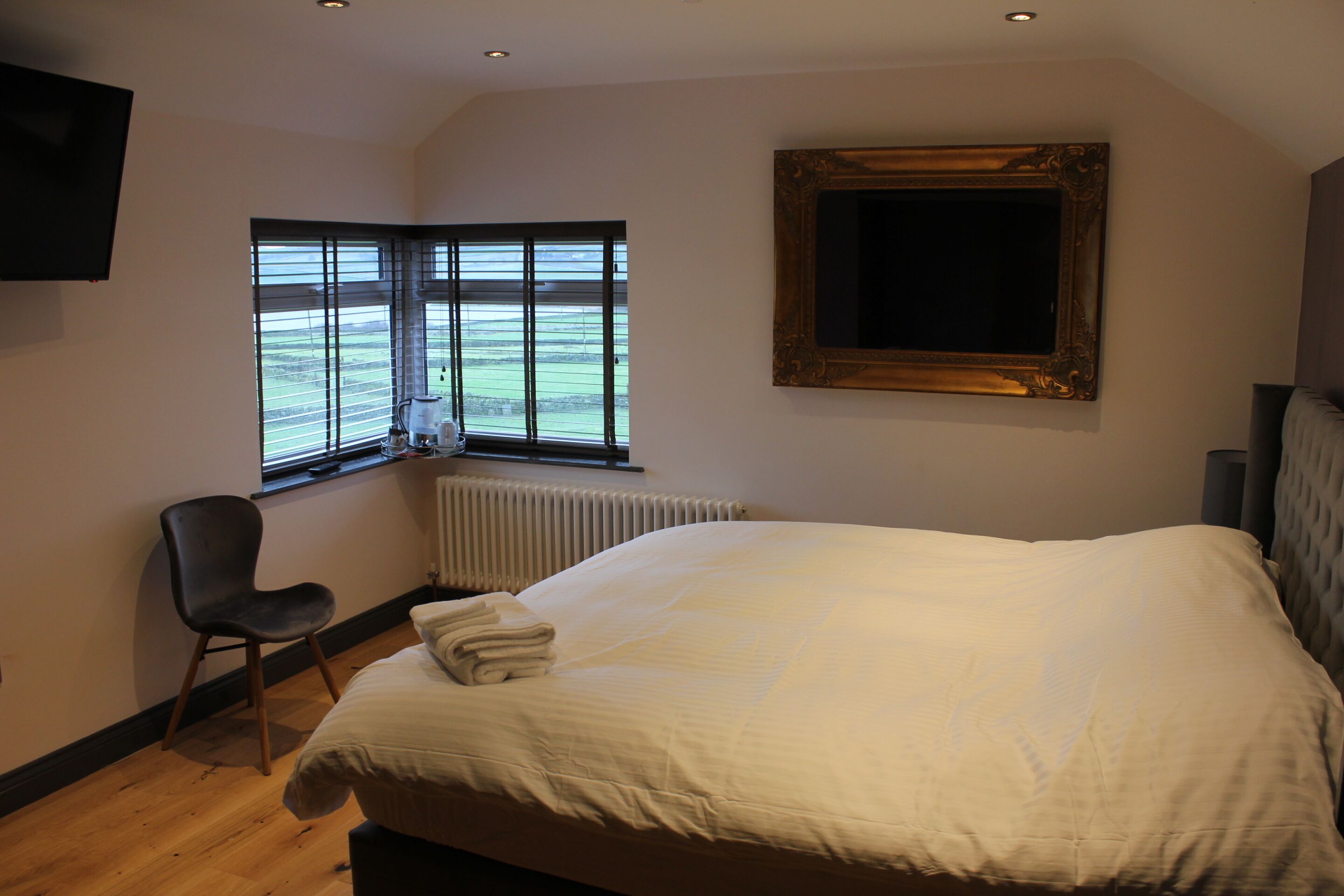 A bedroom with a large bed, a chair, a window with blinds, a radiator, and a television mounted on the wall.