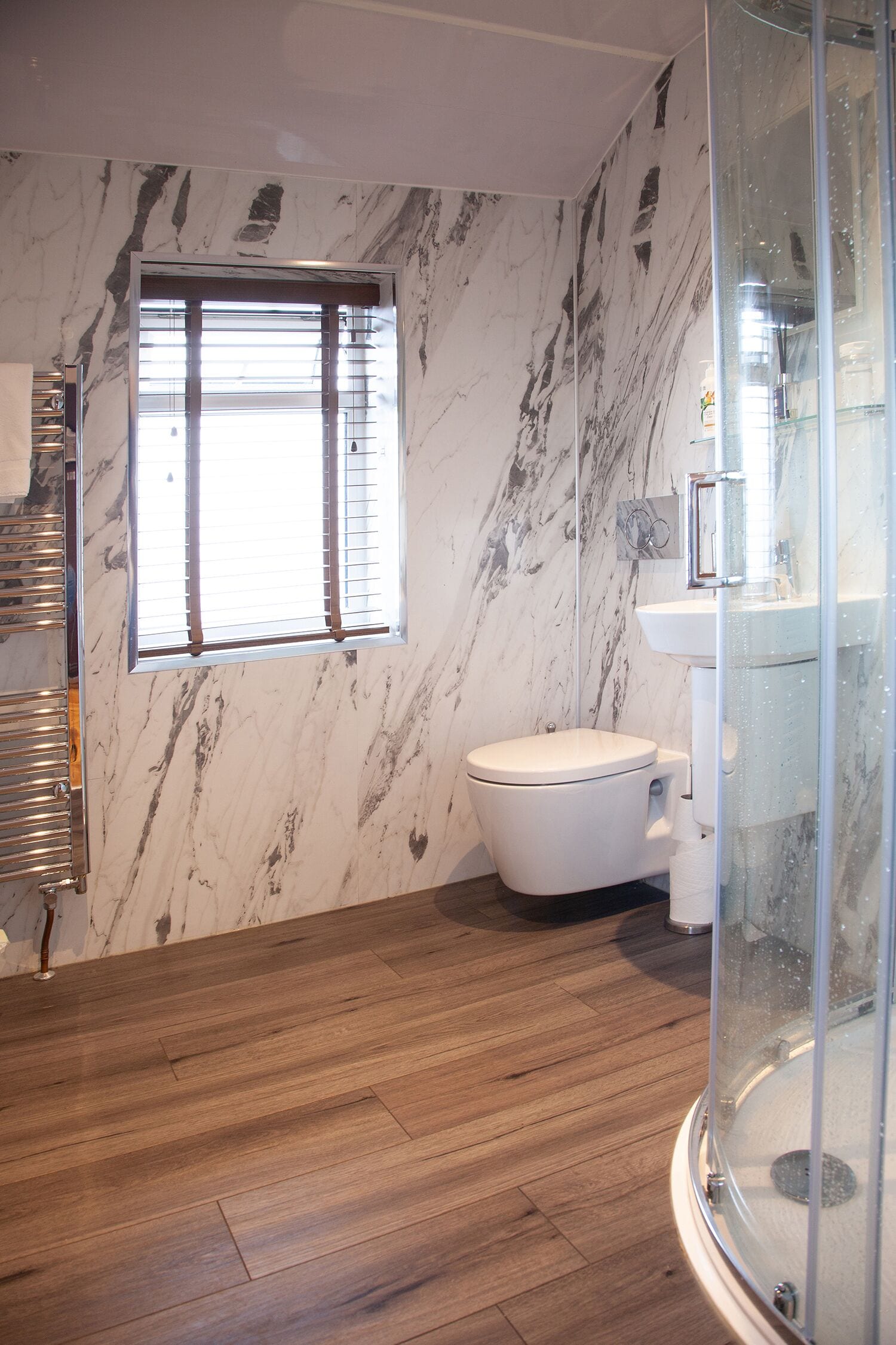 A bathroom with a marble wall, a toilet, a glass shower enclosure, and a wooden floor.
