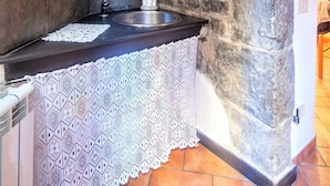 Une cuisine avec un mur en pierre, un plan de travail noir, un évier blanc et une nappe à motifs.