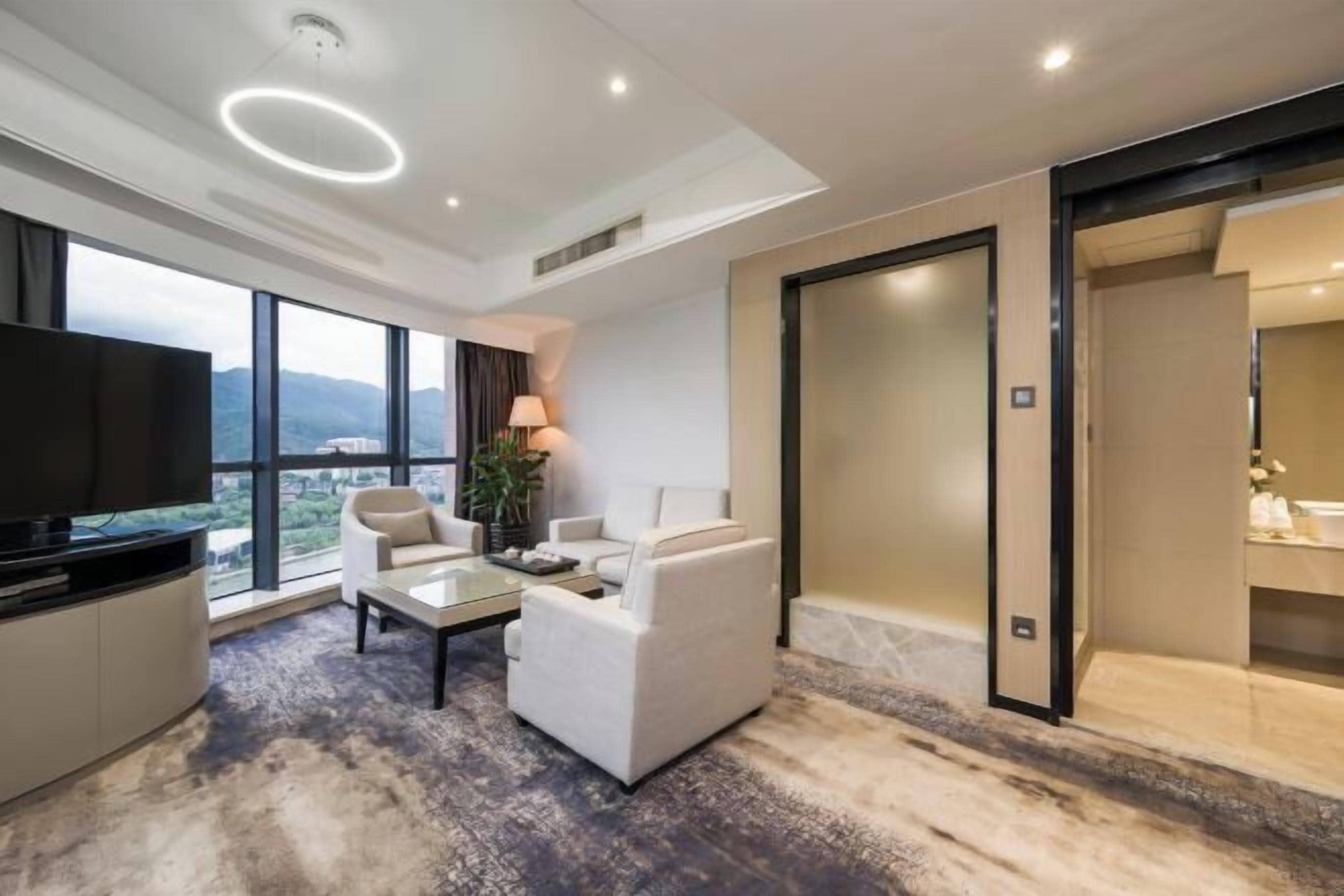 A modern hotel room with a large TV, comfortable seating, a glass coffee table, and a view of the mountains.