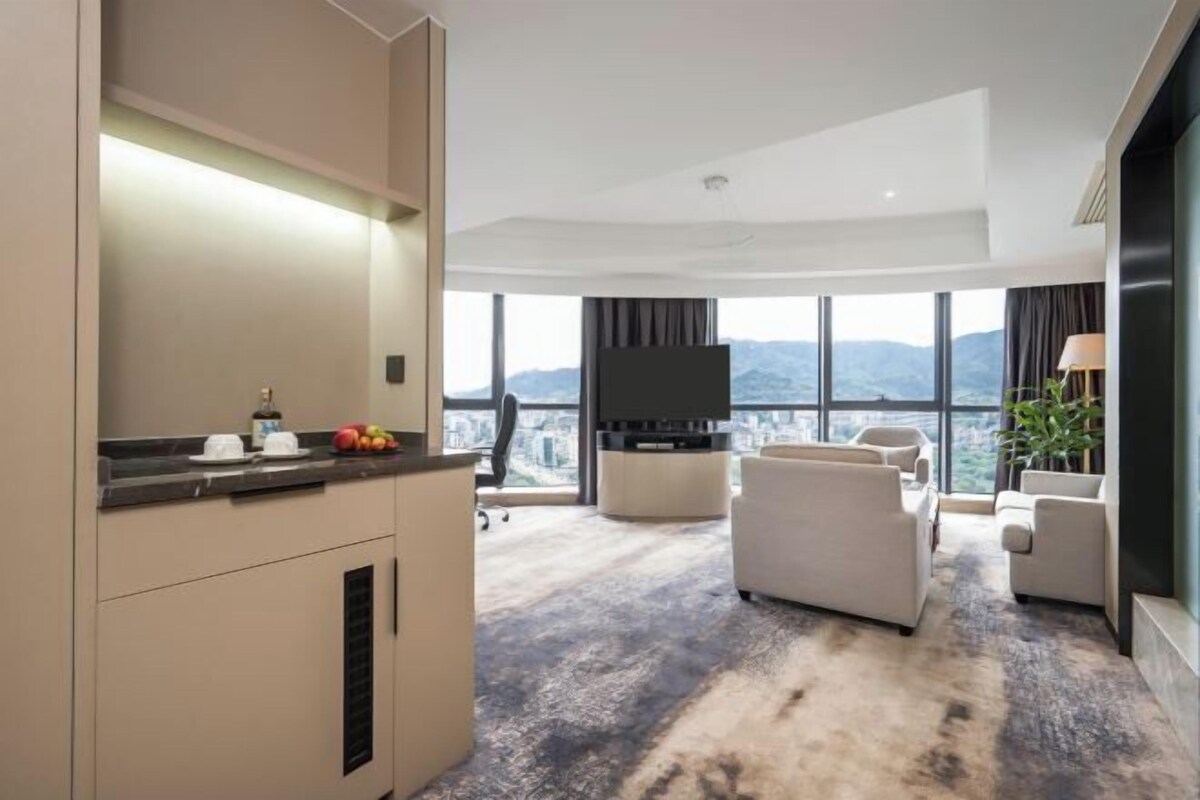 A modern hotel room with a kitchen area, a seating area with a sofa and armchairs, a flat-screen TV, and a large window offering a city and mountain view.
