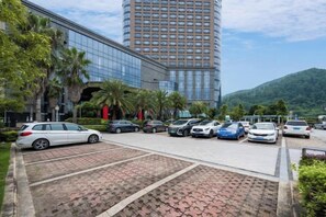 A parking lot with several cars, a modern building with glass facade, and a mountain in the background. - Minzhong Grand Hotel (Youxi)