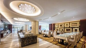 Un'ampia hall d'albergo con pavimento in marmo, una reception centrale e una sala da pranzo con tavoli e sedie rotonde.