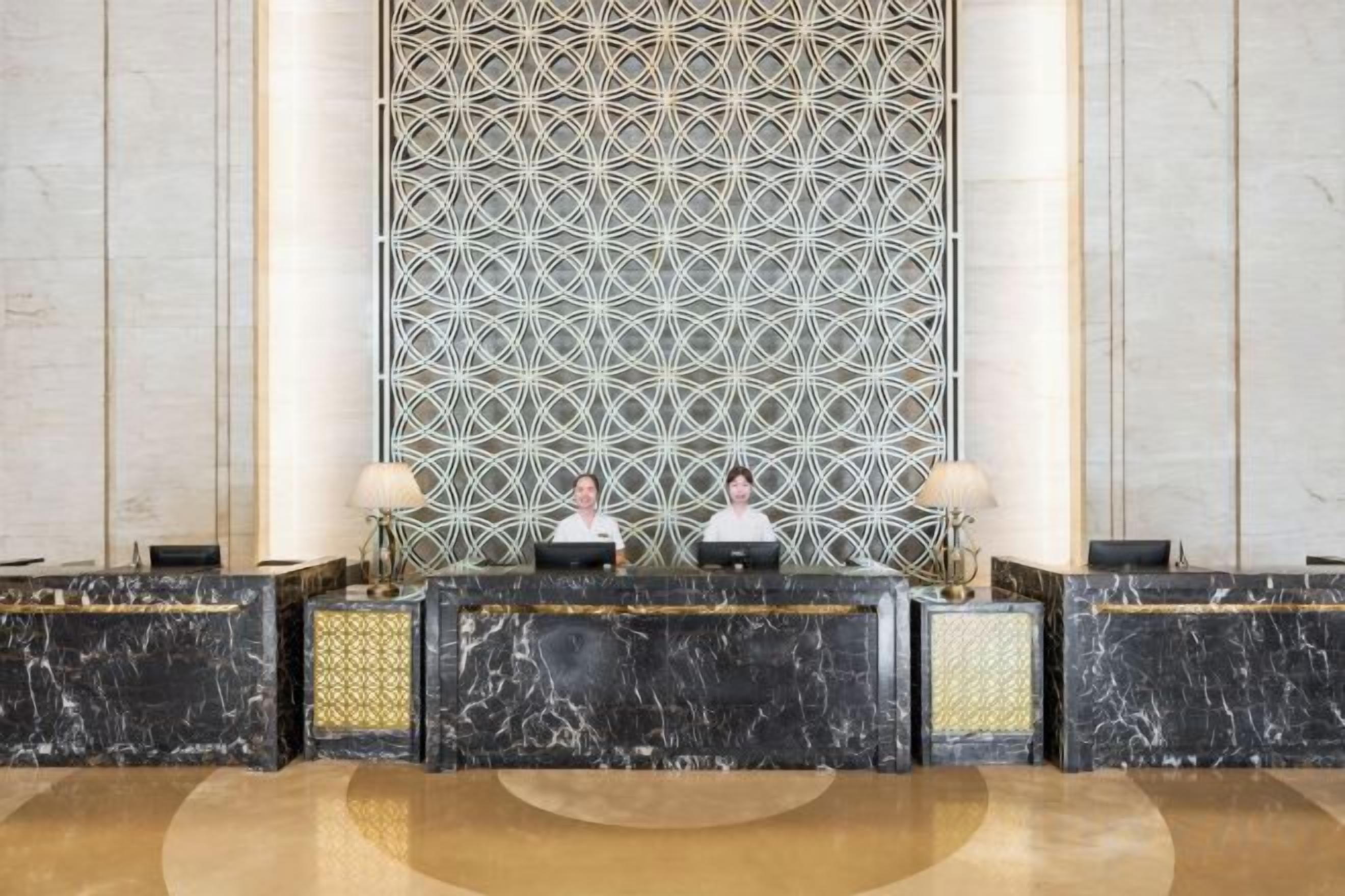 A reception area with a marble reception desk, two receptionists, and a patterned wall behind them.