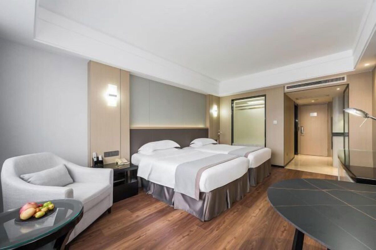 A hotel room with two beds, a sofa, a small table with fruit, and a desk with a lamp.