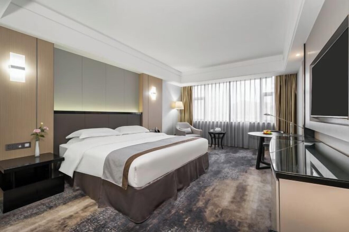 A modern hotel room with a large bed, a desk, a TV, and a window with curtains.