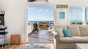 Ein gemütliches Wohnzimmer mit einem beigen Sofa und einem Couchtisch, eine Holzterrasse mit Grill und Blick auf den Strand.