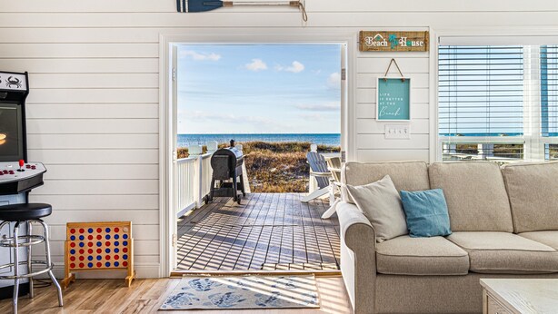 Uma sala de estar aconchegante com um sofá bege e uma mesa de centro, um deck de madeira com churrasqueira e vista para a praia.