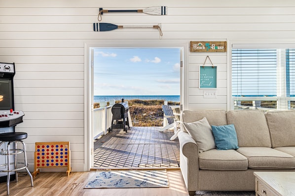 Een gezellige woonkamer met een beige bank en een bijzettafeltje, een houten terras met een barbecue, en uitzicht op het strand.