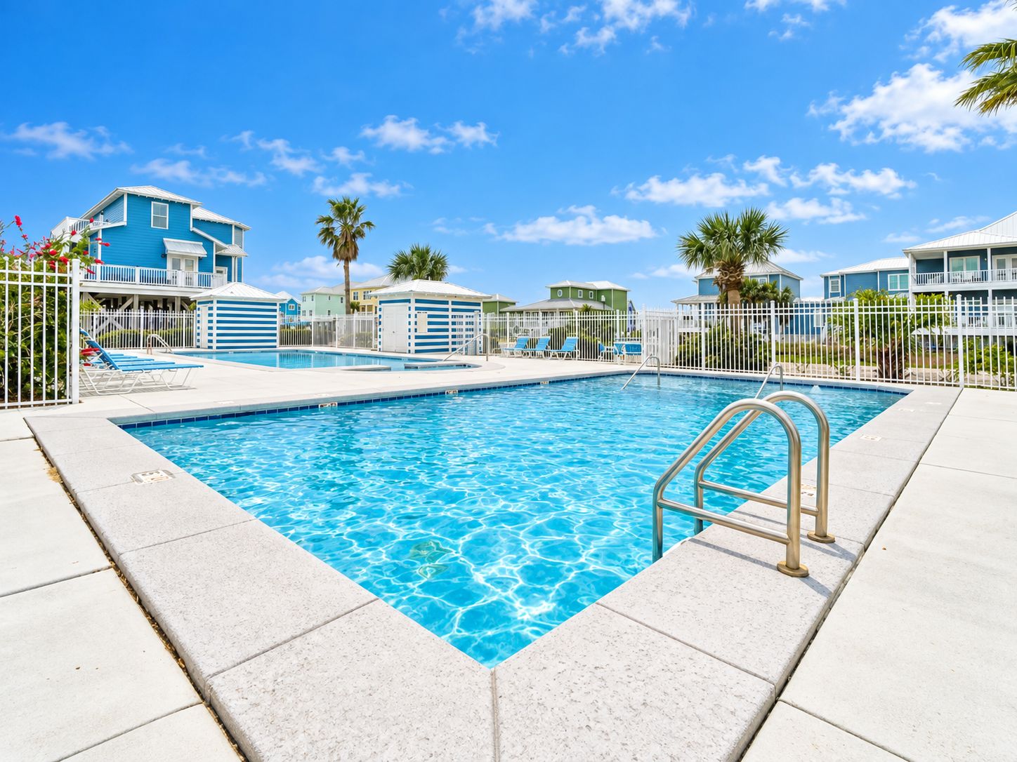 A swimming pool with a metal ladder, surrounded by a white fence, with buildings in the background.