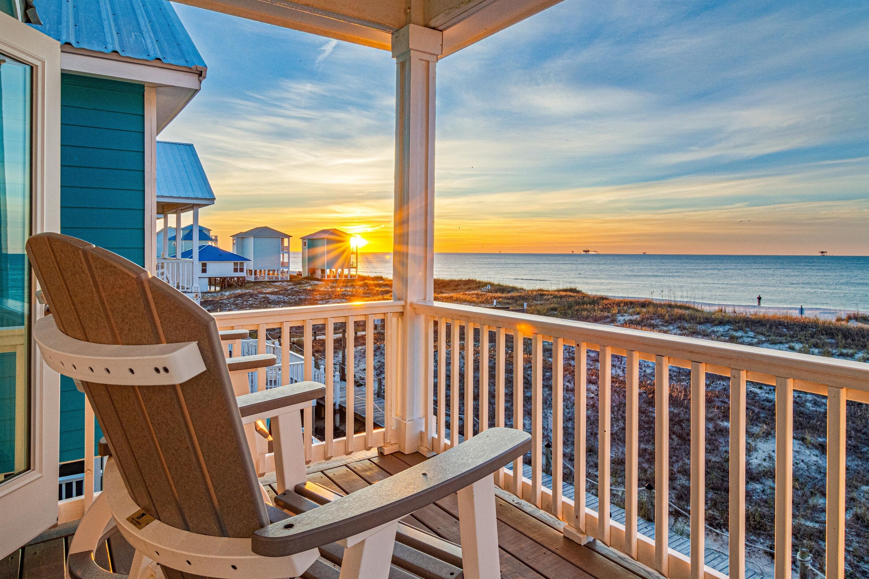 A wooden deck with a chair, overlooking a beach with a sunset.