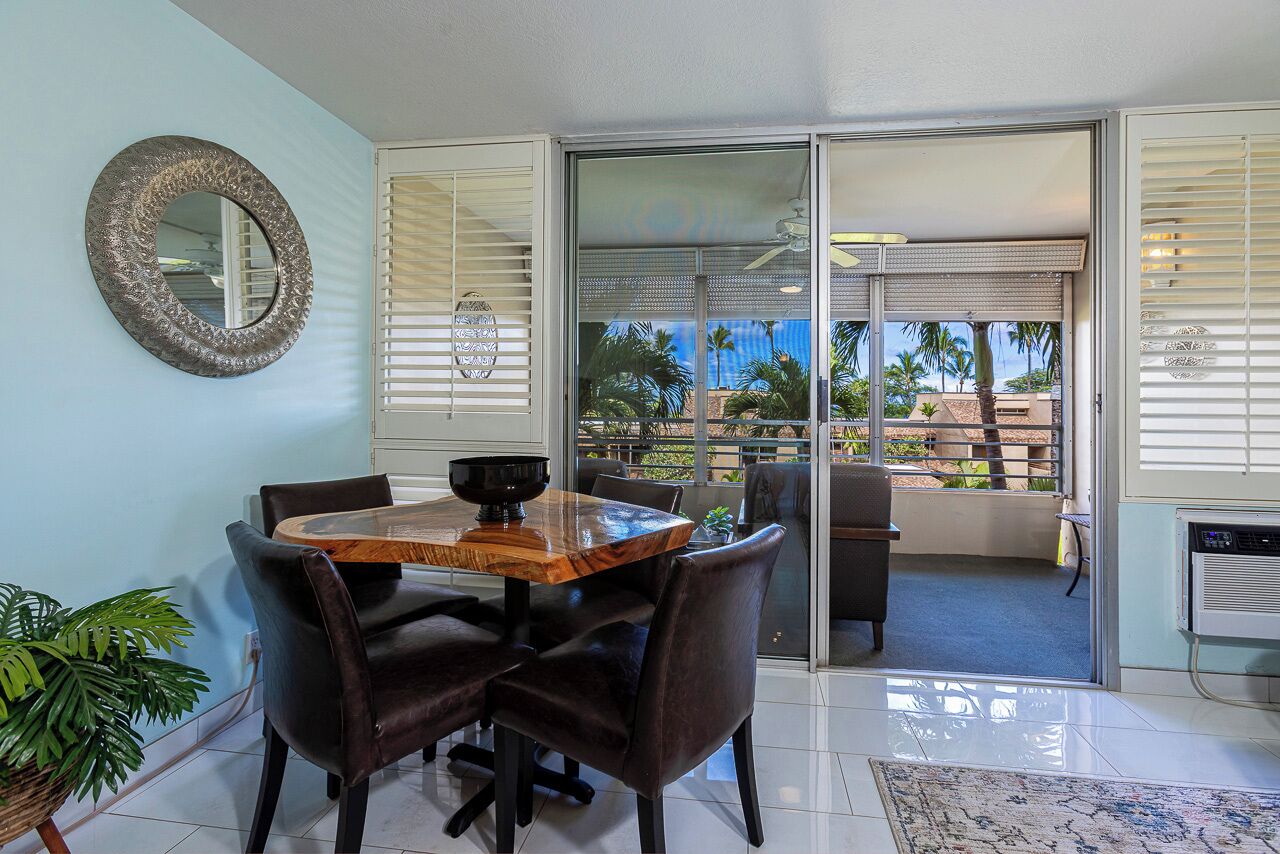 A dining area with a wooden table and chairs, a large mirror, and a view through a sliding glass door to an outdoor patio with a ceiling fan.