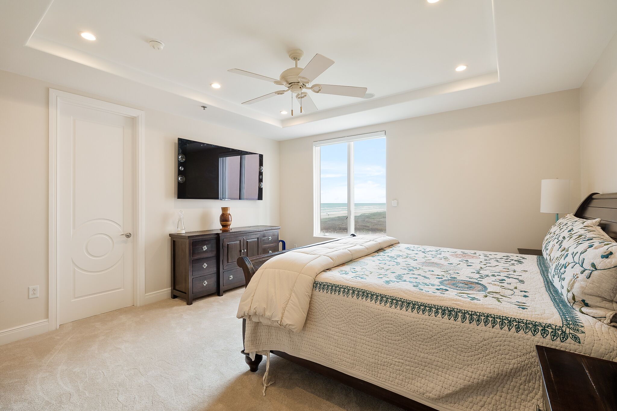 Une chambre à coucher comprenant un lit, un ventilateur de plafond, un téléviseur fixé au mur, une fenêtre et une commode.