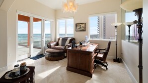 A spacious living room with a large window, a chandelier, a desk, and a leather sofa.