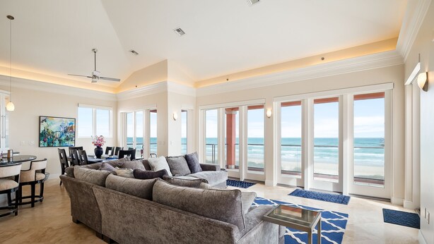 A spacious living room with a large gray sofa, a dining area with a round table and chairs, and large windows offering ocean views.