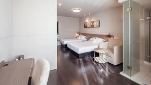 A modern hotel room with a wooden desk, two beds, a glass shower enclosure, and a framed picture on the wall.