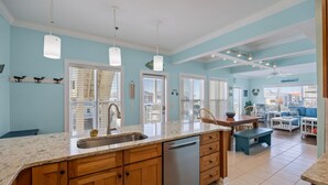 A kitchen with a large island, wooden cabinets, and a dishwasher. There are multiple windows with blinds, a ceiling fan, and a dining area with a table and chairs. - CottageVacations4u "Shore Thing"- ocean views-direct beach access-pool club<br>Golf cart friendly (Ocean Isle Beach)