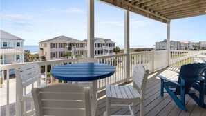 A wooden deck with a round table and chairs, overlooking a beachfront with multiple buildings. - CottageVacations4u "Shore Thing"- ocean views-direct beach access-pool club<br>Golf cart friendly (Ocean Isle Beach)