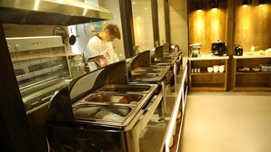 Montaje de bufé con múltiples recipientes de acero inoxidable, una persona con vestimenta de chef y una barra con equipo de cocina.
