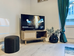 A modern living room with a flat-screen TV on a wooden stand, a black ottoman, a vase with greenery, and a window with curtains.