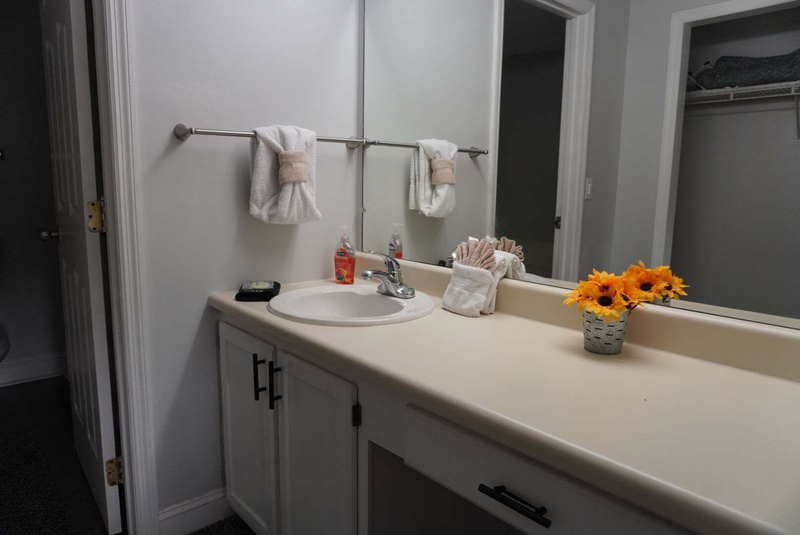 Un baño con un lavamanos blanco, un espejo, un perchero con toallas, un pequeño florero con flores y una puerta.